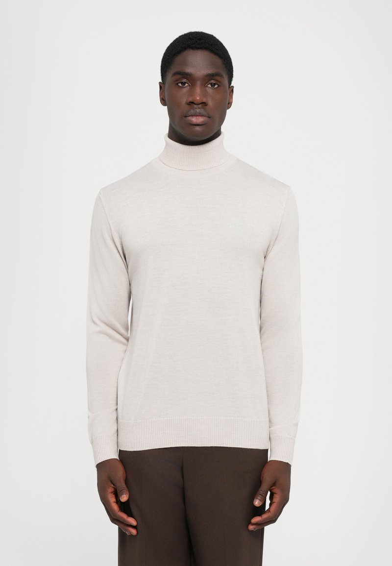 Uomo che indossa un maglione a collo alto beige chiaro e pantaloni marrone scuro, in piedi contro uno sfondo bianco semplice, rivolto in avanti.