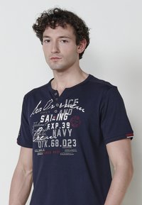 Navy zila īsām piedurknēm krekls ar Henley apkakli, kas izceltas ar baltiem un sarkaniem grafiskajiem drukājumiem, pogām un svītrām uz piedurknēm.