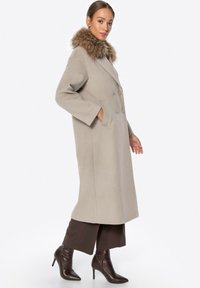 Beiger, doppelt geknöpfter Mantel mit Pelzkragen, lange Ärmel und Seitentaschen, kombiniert mit braunen Hosen und Stiefeletten mit Absatz.