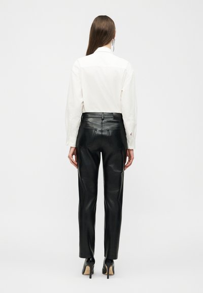 Mulher de costas, vestindo uma camisa branca de manga comprida, calças pretas de couro de cintura alta e sapatos pretos de salto alto.