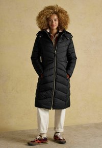 Melnā pūkainā jaka ar kapuci, ar zigzaga kvadrātu rakstu un zelta rāvējslēdzēju. Apvienota ar baltiem biksēm un brūnām kedām.