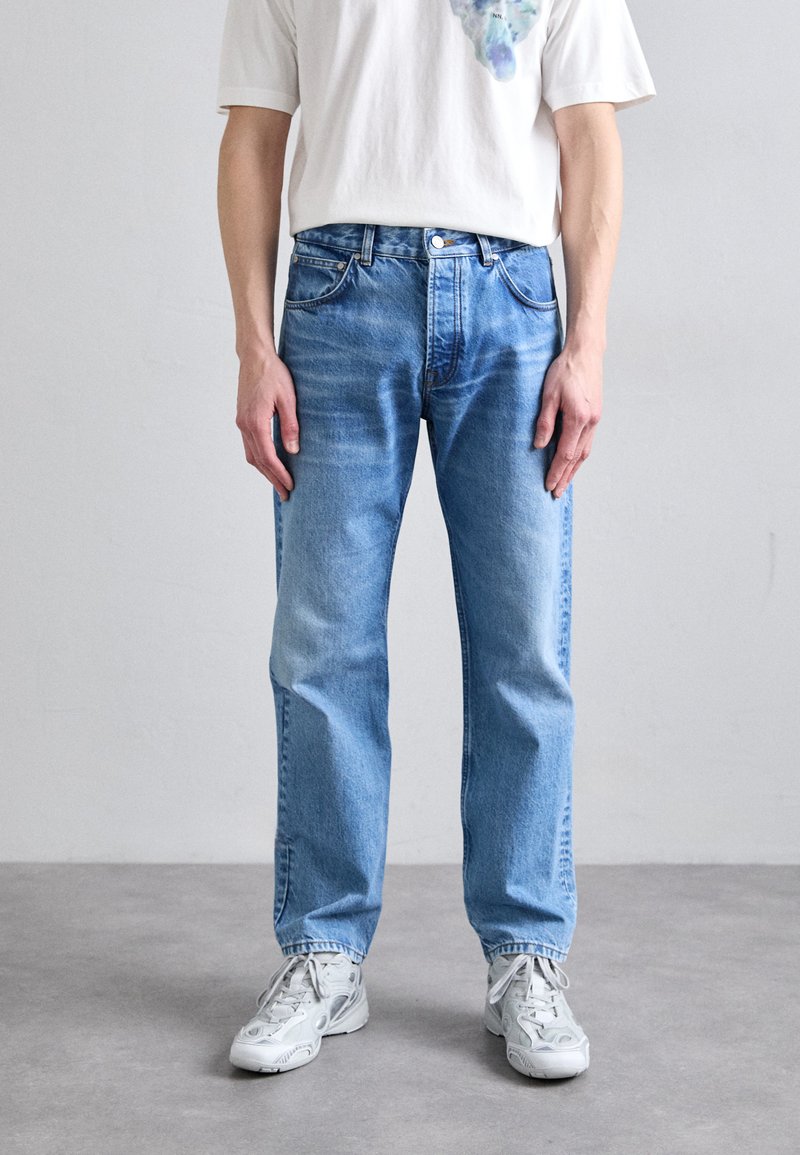 Jeans de mezclilla azul claro con corte recto, un ligero desgaste, cinco bolsillos y cierre de botón. Llevados con una camiseta blanca y zapatillas ligeras.