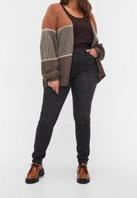 Cárdigan de punto marrón y gris con mangas sueltas, superpuesto a una camiseta de tirantes oscura; combinado con jeans oscuros y zapatos marrones con cordones.