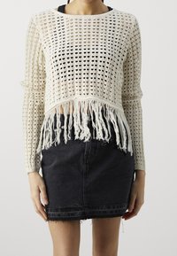 Kremowy, szydełkowy top w stylu crop z długimi rękawami i frędzlami na dole, z luźnym wzorem dzianiny. W połączeniu z czarną spódnicą z wysokim stanem z denimu.