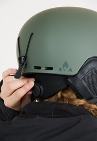 Groene matte skihjelm met ventilatiegaten, verstelbare riem en padding. Heeft zwarte accenten en een logo aan de zijkant. Persoon past de pasvorm aan.