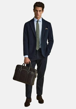 Mies tummansininen bleiseri, vaaleansininen paita, vihreä solmio, tummat housut, ruskeat kengät, pitää tummanruskeaa nahkaista salkkua, jossa on kaksi kahvaa.