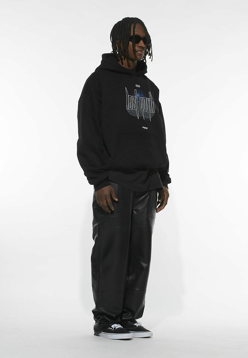 Sudadera negra con estampado gráfico en el pecho, combinada con pantalones de cuero sintético negros; el modelo lleva gafas de sol negras y zapatillas negras.