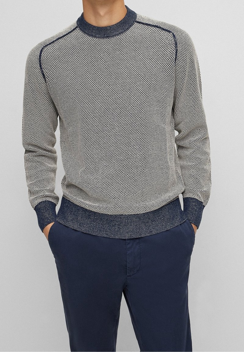 Homme portant un pull en maille gris texturé et bleu marine avec un pantalon bleu marine, debout les mains dans les poches contre un fond uni.