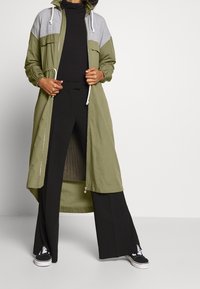 Chaquetón largo verde olivo con panel superior gris, capucha ajustable, dos bolsillos en el pecho y un zipper frontal. Combinado con pantalones de pierna ancha negros y zapatillas.