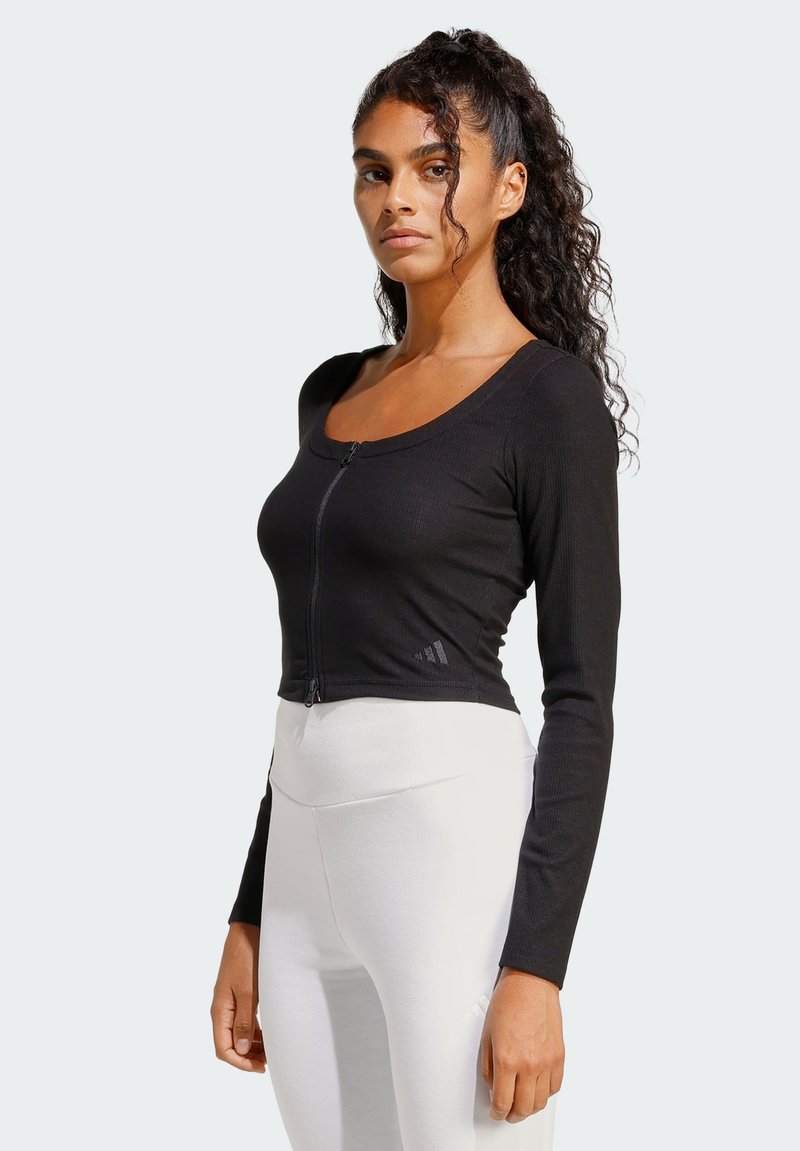 Woman with long curly hair wearing a black zip-up long sleeve top and high-waisted white leggings standing against a plain background.