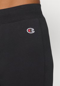 Melna kokvilnas sporta bikses ar elastīgu jostasvietu un mazu izšūtu logotipu sarkanā, baltā un melnā krāsā, kas novietots kreisajā augšstilbā.