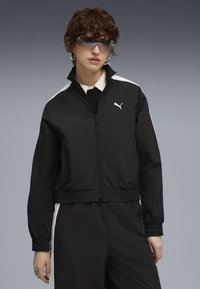 Czarna kurtka zapinana na zamek z elastycznym dołem i mankietami, z białą boczną paskiem i małym białym logo na klatce piersiowej. Lekki materiał.