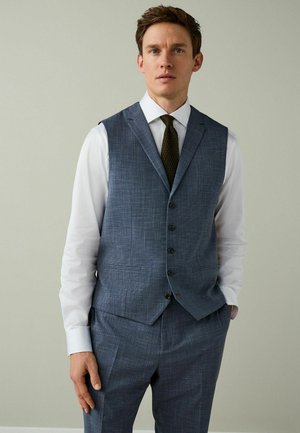 Man wearing blue checkered suit vest and matching trousers, white dress shirt, and dark textured tie, standing with one hand in pocket.