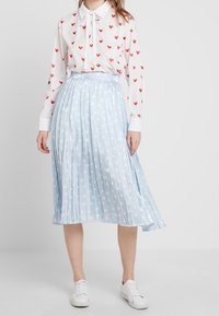 Vit blus med röd hjärtmotiv, matchad med en ljusblå plisserad kjol med vita stjärndetaljer, och vita sneakers.