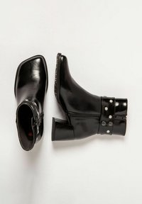 Botas de cuero negras hasta el tobillo con punta cuadrada, tacón cuadrado y cremallera lateral. Incluyen una tira decorativa con tachuelas alrededor del tobillo.