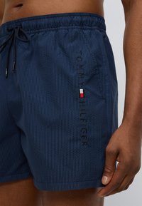 Μπλε ναυτικό μαγιό με υφή υφάσματος, ελαστική μέση και κορδόνι. Διαθέτει κεντημένο λογότυπο "TOMMY HILFIGER" και έγχρωμο διακοσμητικό λεπτομέρεια.