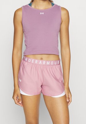 Kobieta w bezrękawnym fioletowym crop topie i różowych spodenkach Under Armour z widocznym logo na pasku, stoi na jednolitym tle.