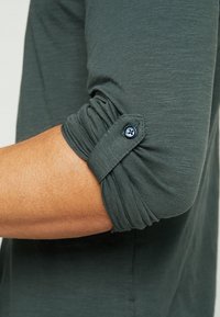 Dunkelgrünes Langarmhemd mit hochgekrempelten Ärmeln, das an der Manschette einen blauen Knopf hat. Der Stoff wirkt weich mit einer strukturierten Oberfläche.
