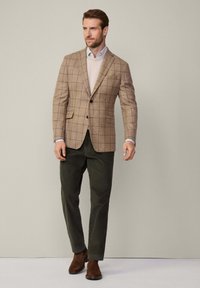 Blazer a scacchi marrone sopra un maglione crema, abbinato a pantaloni verde scuro e stivali in pelle marrone. Tessuto morbido e vestibilità su misura.
