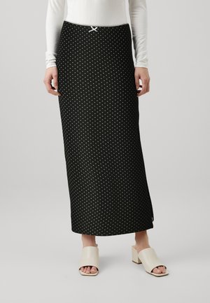 Black knee-length skirt with white polka dots, featuring a waistband with a decorative bow. Worn with beige open-toe block-heeled mules.