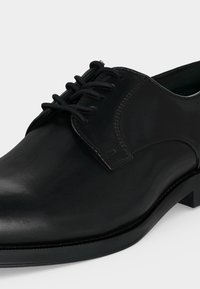 Chaussure de ville en cuir noir avec laçage fermé, talon bas et finition lisse et polie, présentée sur un fond clair et uni.