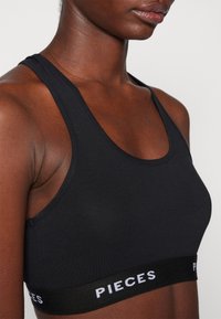 Soutien-gorge de sport noir en tissu lisse et extensible. Il présente une large bande noire avec "PIECES" en lettres blanches, des fines bretelles et un décolleté arrondi.
