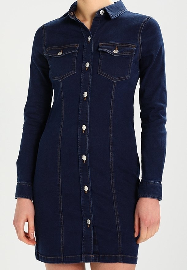 Kvinna iklädd en figurnära mörkblå jeansklänning med långa ärmar, knappstängning fram och två bröstfickor med klaffar.