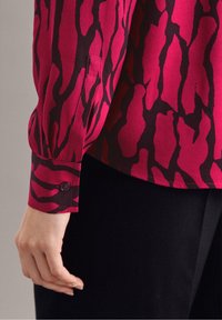 Cuff di una camicia a maniche lunghe con motivo astratto rosso e nero, indossata con pantaloni neri, vista di lato su un braccio di una persona.