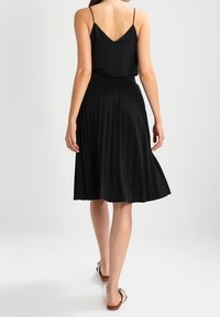 Femme portant une jupe midi plissée noire et un haut à bretelles noires, marchant pieds nus sur un fond blanc.