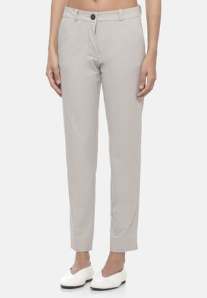 Donna che indossa pantaloni beige chiaro a gamba dritta e scarpe bianche slip-on, in piedi con le braccia rilassate lungo i fianchi contro uno sfondo neutro.