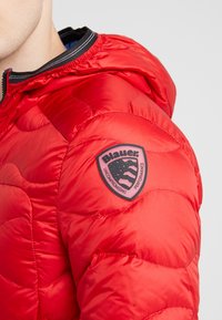 Detailní záběr na rukáv červené prošívané bundy s černou a růžovou nášivkou loga Blauer s nápisem "Neochvějný výkon."