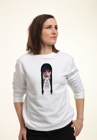 Žena s rovnými hnědými vlasy v bílém mikině, která má na sobě grafiku vážné dívky s copy a text "ZÓNA BEZ OBJÍMÁNÍ."