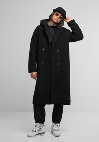 Black double-breasted coat in a plush texture, paired with a black hoodie, black joggers, and white sneakers with teal accents.