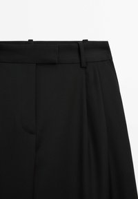 Zwarte op maat gemaakte broeken met riemlussen, verborgen sluiting aan de voorkant en geplooide details aan de rechterkant op een witte achtergrond.