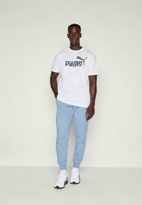 Bela bombažna majica s črnim vojaškim vzorcem in logotipom "PUMA", v kombinaciji s svetlo modrimi trenirka hlačami in belimi športnimi copati s rdečimi detajli.