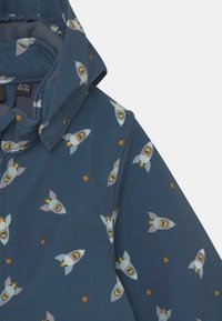 Blaue Kinderjacke für Kleinkinder mit Raketen- und Sternenmuster, Größenetikett zeigt 0-2 Monate, 50/56, abnehmbare Kapuze sichtbar.