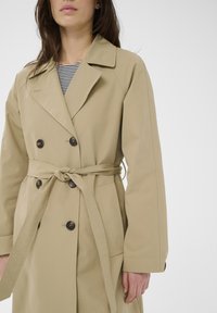 Femme portant un trench-coat beige à double boutonnage avec des boutons noirs et une ceinture nouée à la taille, par-dessus une chemise rayée noire et blanche.