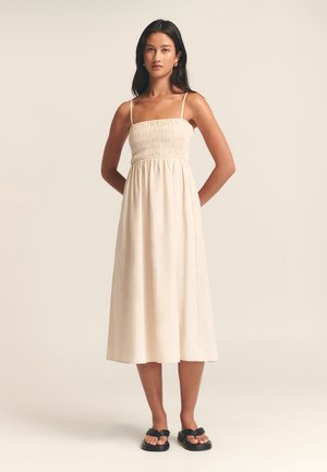 Light beige, knee-length dress with a smocked bodice, thin straps, and a flowy skirt. Worn with black flat sandals.