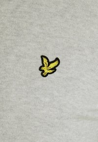 Lichtgrijze stof heeft een klein zwart en geel geborduurd vogellogo, dat gedetailleerde vleugels en klauwen toont tegen de gestructureerde achtergrond.