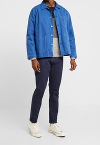 Blaue Denimjacke mit zwei Fronttaschen, Knopfverschluss und Kragen, kombiniert mit einem grauen Pullover und navyblauen Hosen, abgerundet mit weißen Sneakers.