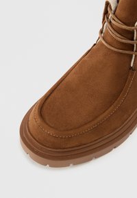 Brun ruskindsstøvle med en afrundet tå, struktureret overflade og syede detaljer. Har en gummisål og snørebånd omkring anklen.