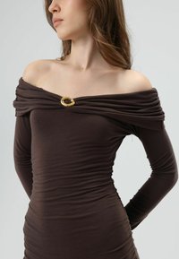 Bruine jurk met blote schouders en lange mouwen, met geplooide textuur en een gouden ronde accent aan de halslijn. Gladde, rekbare stof.