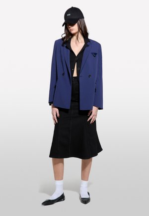 Blazer doppiopetto blu marino, top corto nero, gonna di jeans nera, calzini bianchi e ballerine nere a punta. Cappellino con logo del marchio.