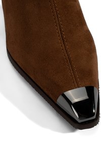 Braune Wildleder-Ankle-Boots mit spitzer Zehe, ausgestattet mit einer glänzenden schwarz-metallischen Zehenschutzkappe und sichtbaren Nähdetails auf dem Obermaterial.