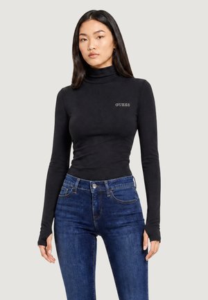 Mujer con cabello largo y oscuro que lleva un top negro de manga larga de cuello alto de Guess y vaqueros azul de cintura alta sobre un fondo liso.