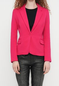 Femme portant un blazer ajusté rose vif sur une chemise noire et un pantalon noir texturé, les mains détendues le long du corps.