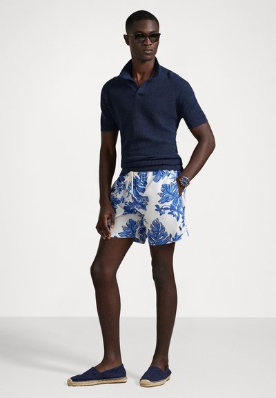 Jūras spēku šūtas polo krekls, baltas šorti ar ziliem tropiskiem lapu rakstiem, ikdienas espadrilu apavi. Aksesuāri ietver tumšas saulesbrilles un pulksteni.