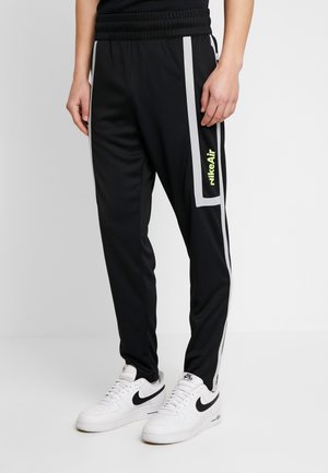 Personne portant un pantalon de survêtement Nike Air noir avec des bandes blanches sur les côtés et des baskets Nike blanches avec des logos swoosh noirs.