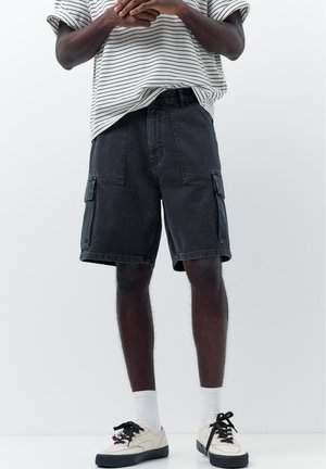 Person iført sorte cargoshorts, hvide sokker, beige sneakers og en hvid stribet skjorte med opkørte ærmer, stående med hænderne foldet.