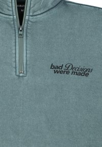 Mintgroene zip-up sweatshirt gemaakt van zachte stof, met een hoge kraag en geborduurde tekst "slechte beslissingen zijn genomen" in het zwart.